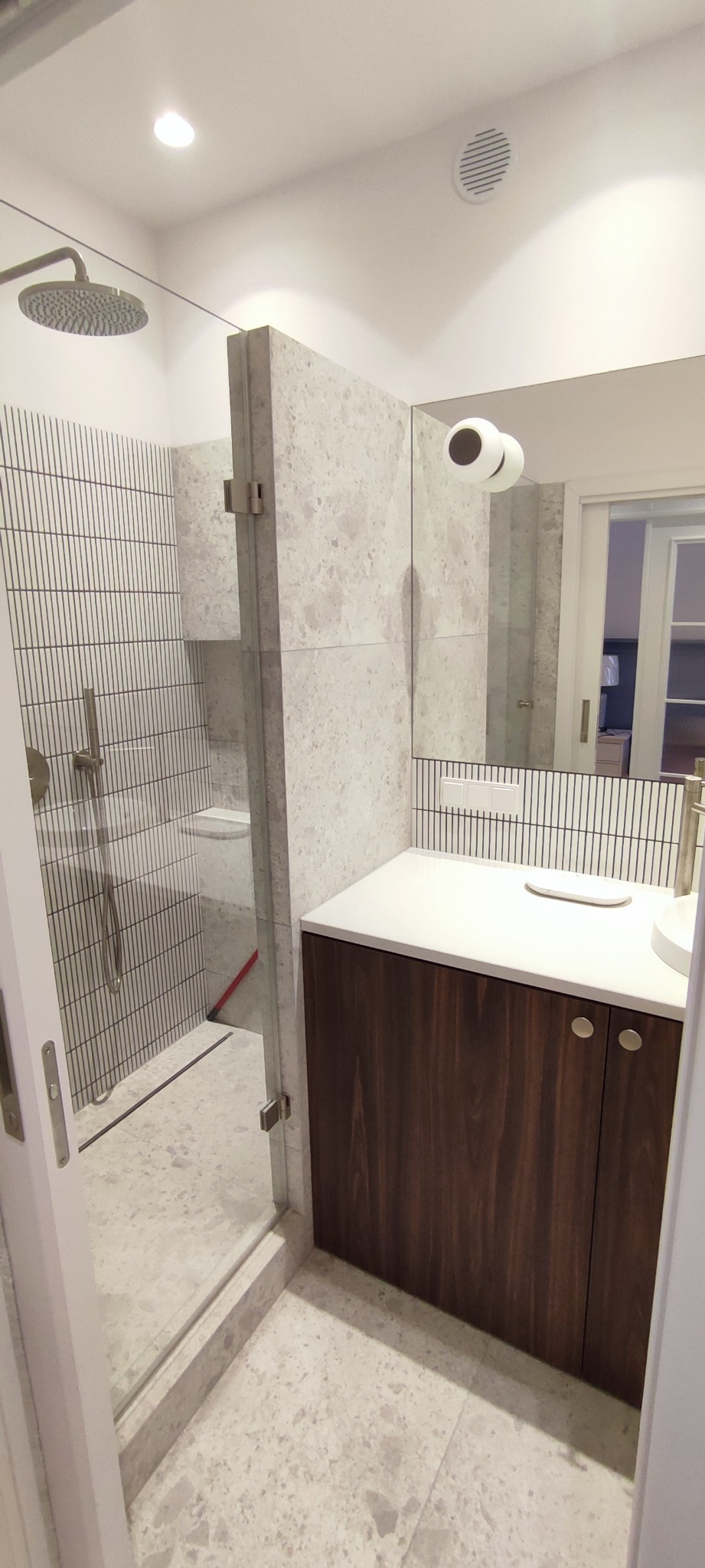 Nowoczesna łazienka z kabiną prysznicową typu walk-in, wykończona płytkami w odcieniach szarości z ciemnymi fugami. Widoczna umywalka nablatowa z szafką z ciemnego drewna i okrągłym lustrem...