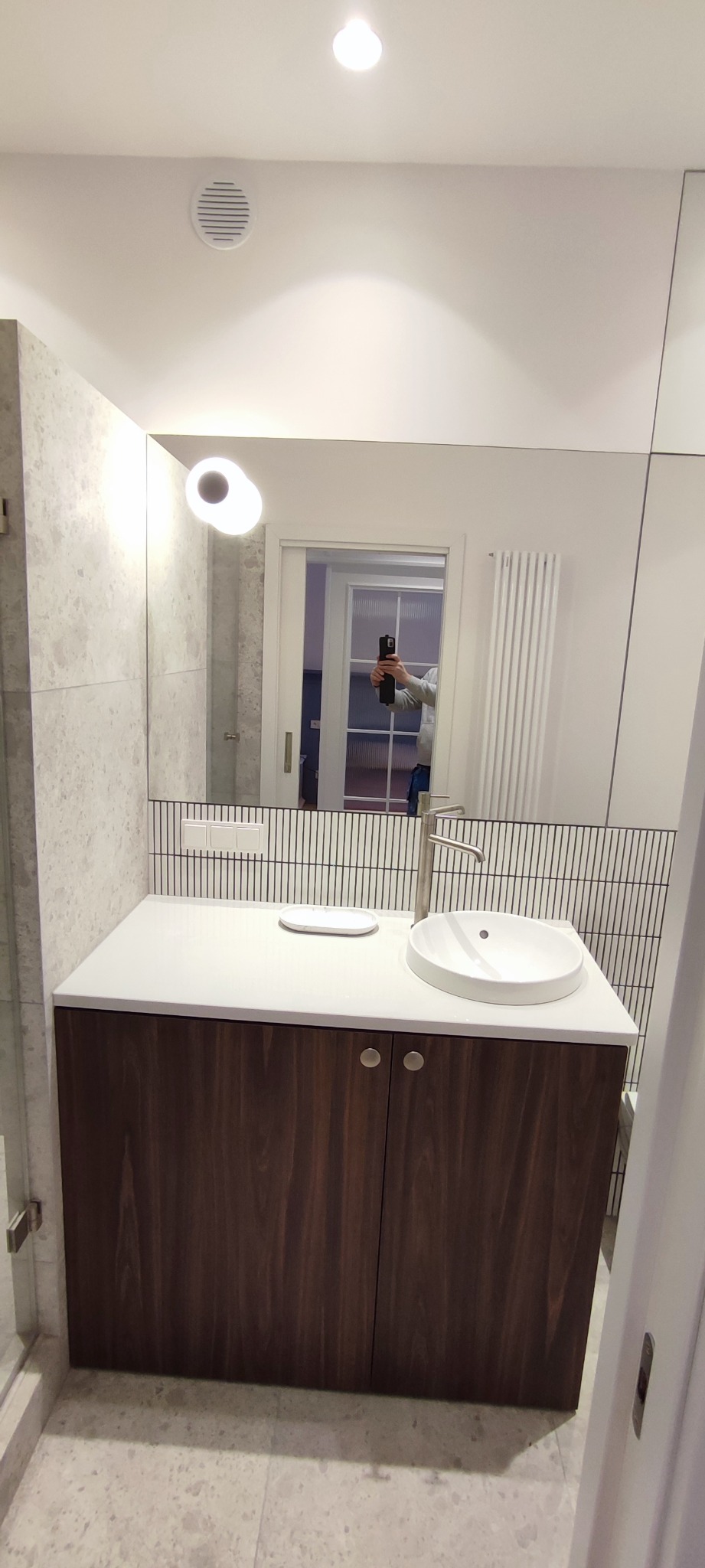 Nowoczesna łazienka z umywalką nablatową, szafką z ciemnego drewna i dużym lustrem, w którym odbija się osoba robiąca zdjęcie telefonem.