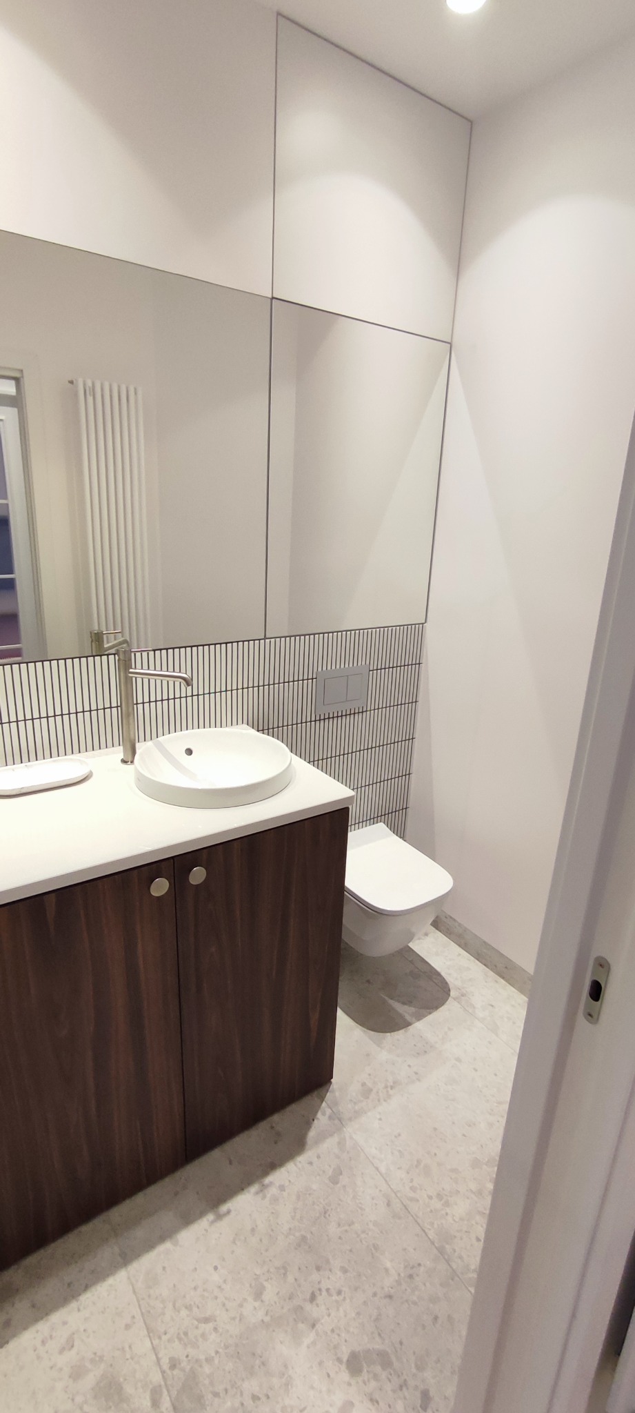 Nowoczesna łazienka z umywalką nablatową, szafką z ciemnego drewna, podwieszaną toaletą i lustrem zajmującym całą ścianę, wykończona płytkami w pionowe paski za toaletą i umywalką.