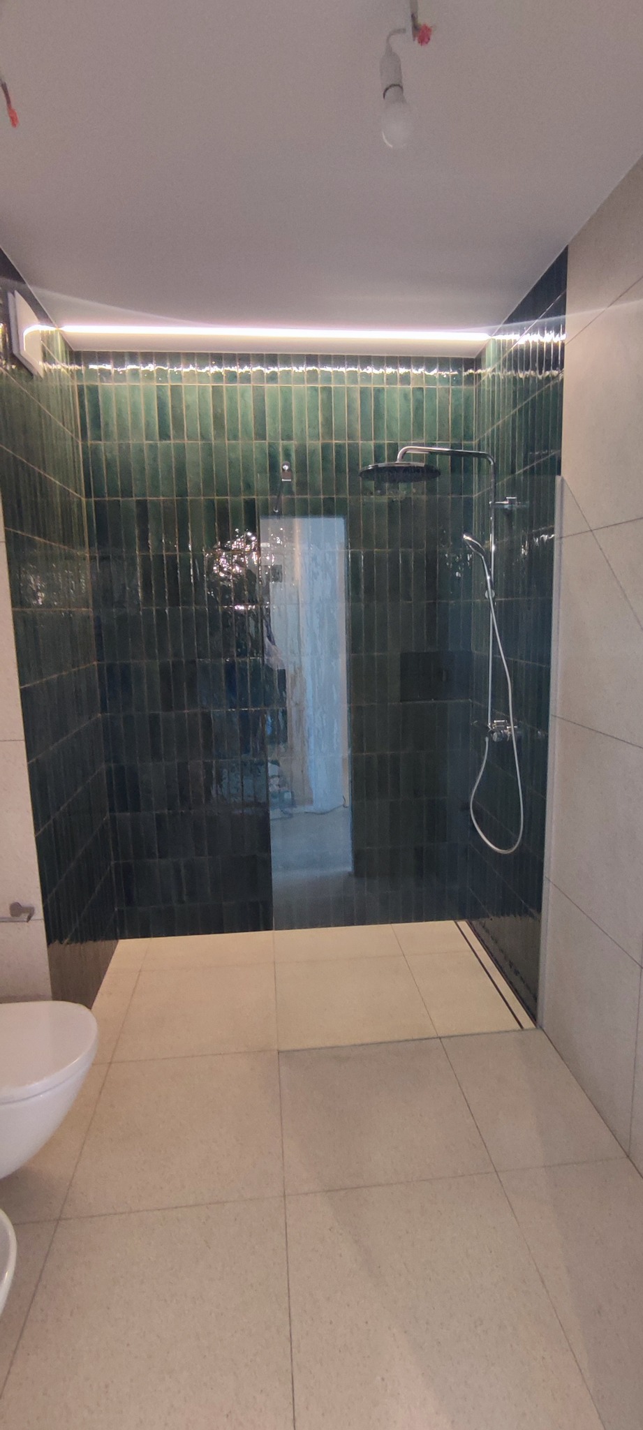 Nowoczesna kabina prysznicowa z ciemnozielonymi, pionowo ułożonymi płytkami, podświetlona listwą LED, widoczne elementy armatury i jasna, kwadratowa podłoga. W tle widoczne pomieszczenie w trakcie...