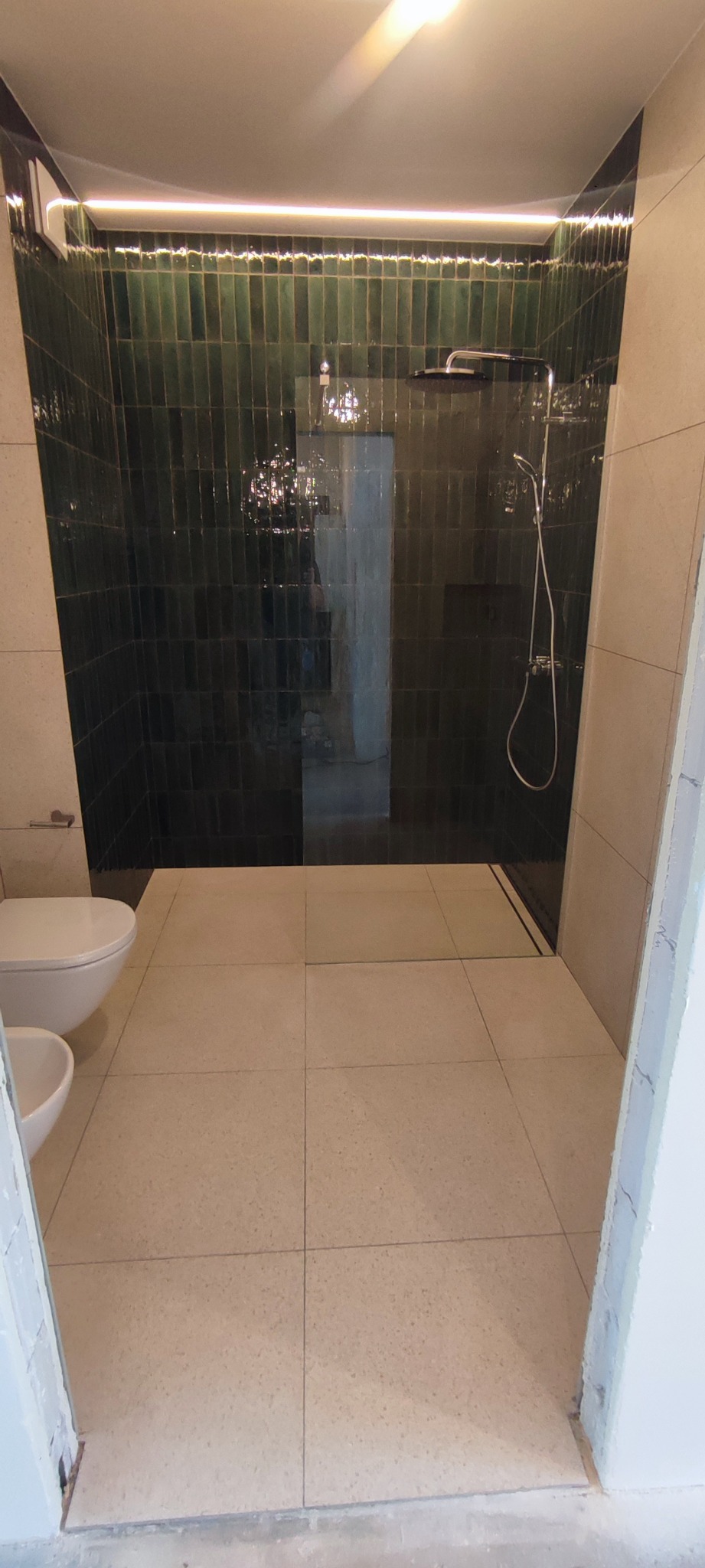 Nowoczesna łazienka z kabiną prysznicową typu walk-in, wyłożona pionowymi, ciemnozielonymi płytkami, oświetlona listwą LED pod sufitem, z widoczną toaletą i bidetem, oraz beżową podłogą z dużych...