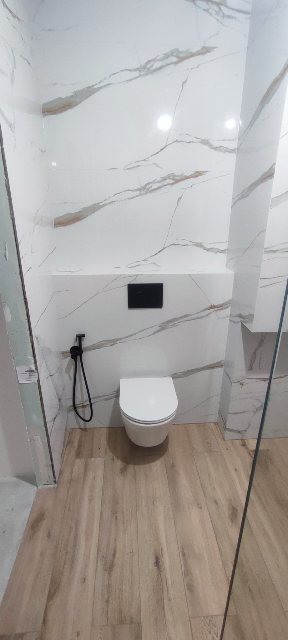 Nowoczesna toaleta podwieszana z czarnym panelem sterującym i bidetką w łazience wykończonej białymi płytkami imitującymi marmur i panelami podłogowymi w kolorze jasnego drewna.