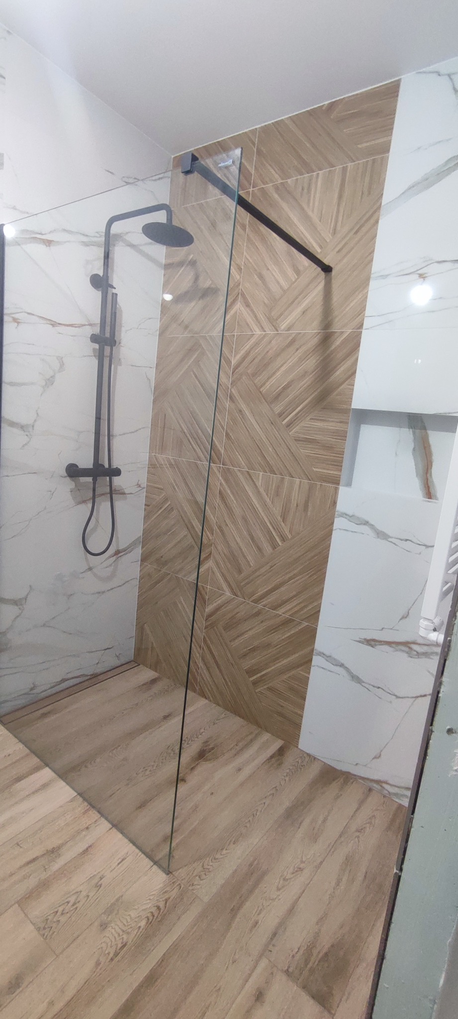 Nowoczesna kabina prysznicowa z czarną armaturą, szklaną ścianką i drewnopodobnymi płytkami na podłodze i części ściany, połączonymi z białymi płytkami imitującymi marmur.