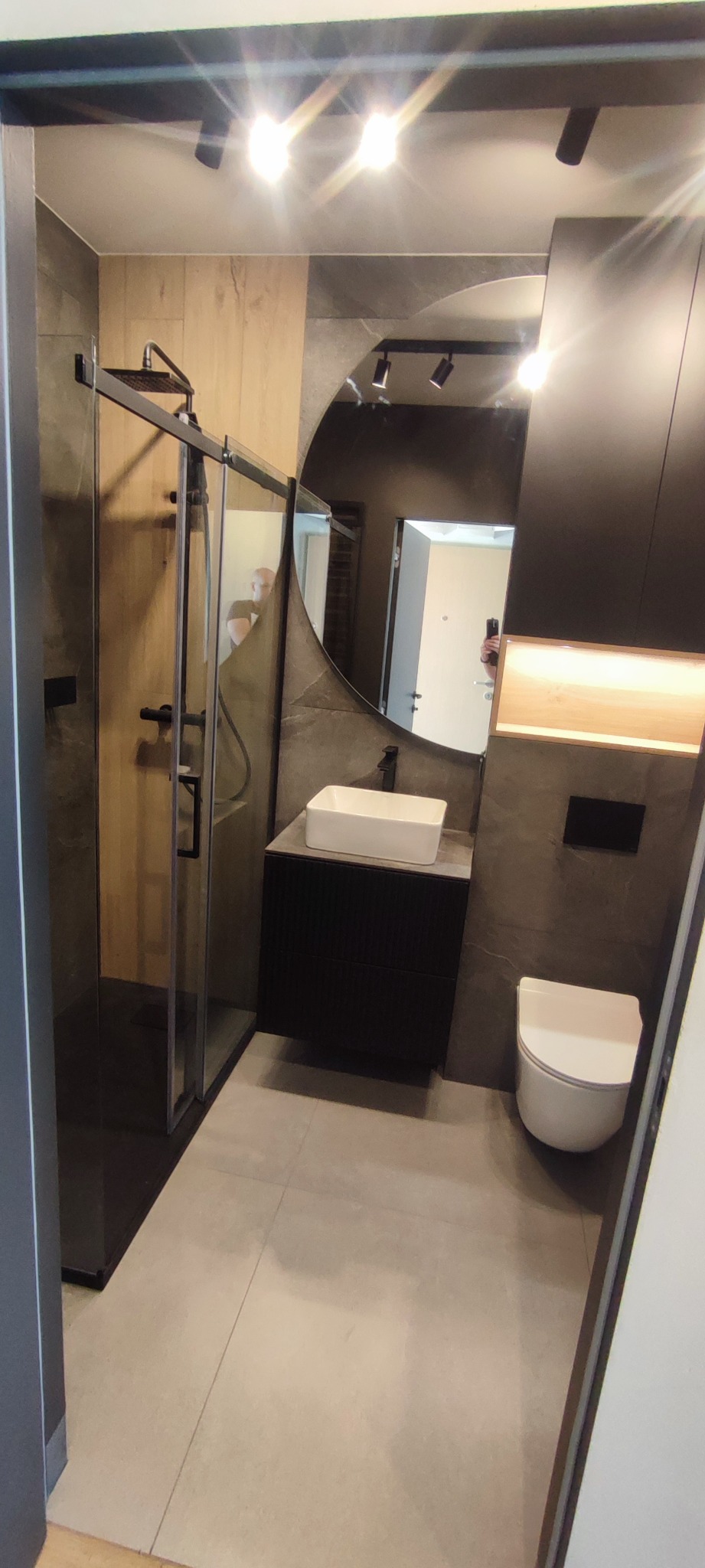 Nowoczesna łazienka z kabiną prysznicową typu walk-in, okrągłym lustrem i czarną armaturą na tle szarych płytek imitujących kamień, oświetlona punktowymi lampami sufitowymi.