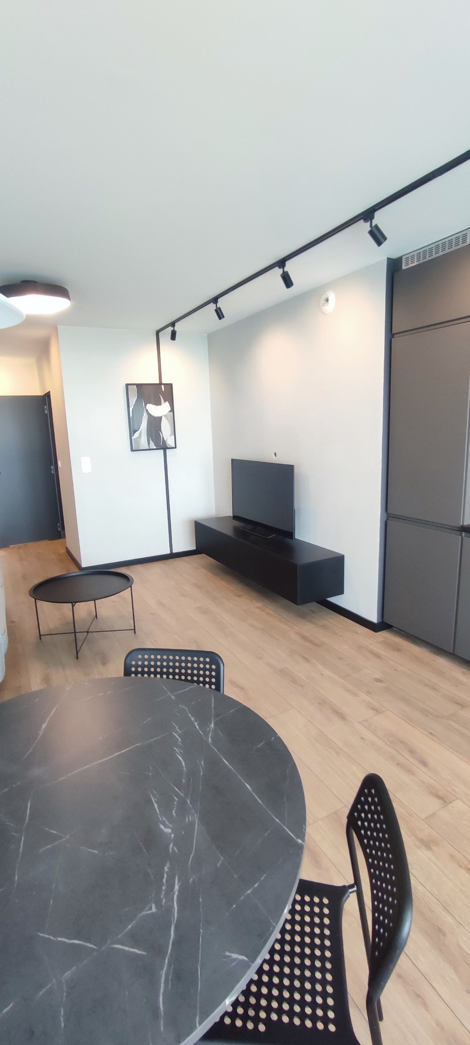 Nowoczesne wnętrze mieszkania z okrągłym stołem z czarnym blatem imitującym marmur, czarnymi krzesłami, podłogą z paneli, czarną szafką RTV i telewizorem, oświetleniem szynowym i szarą zabudową...