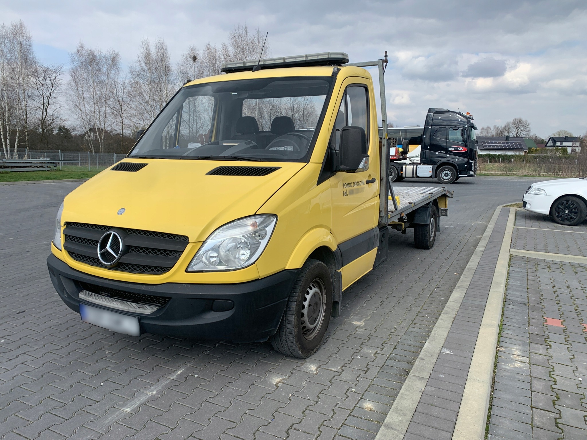Żółty samochód pomocy drogowej Mercedes Sprinter z widocznym napisem 'Pomoc Drogowa' na boku, stojący na parkingu z kostki brukowej, w tle inne pojazdy i drzewa bez liści.