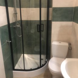 Firma Budowlana Tomasz Jędroszka - Ukończona łazienka z kabiną prysznicową z czarnymi ramami, toaletą i umywalką, wykończona płytkami w pasy zieleni i bieli.