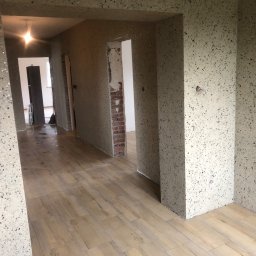 Firma Budowlana Tomasz Jędroszka - Korytarz w trakcie remontu z wykończonymi ścianami w dekoracyjnym tynku z drobnymi kamyczkami, drewnopodobną podłogą i widocznym fragmentem ściany z cegły w przejściu.