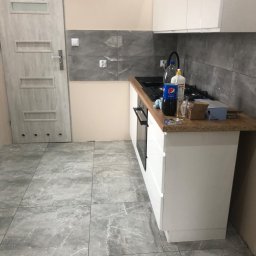 Firma Budowlana Tomasz Jędroszka - Wykończona kuchnia z szarymi płytkami podłogowymi imitującymi marmur, białymi szafkami kuchennymi i szarymi płytkami ściennymi; na blacie widoczne butelki, naczynia i paczka papierosów.