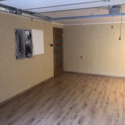 Firma Budowlana Tomasz Jędroszka - Wykończone wnętrze garażu z położoną podłogą z paneli imitujących drewno, widoczne kable wystające ze ściany oraz szyny do bramy garażowej pod sufitem.