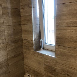 Firma Budowlana Tomasz Jędroszka - Wykończona ściana w łazience z płytek imitujących trawertyn, okno z białymi ramami i tubka silikonu na parapecie, widoczny fragment instalacji wodnej.