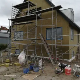 Dom w trakcie ocieplania, ściany pokryte żółtą izolacją, rusztowania dookoła, widoczne narzędzia i materiały budowlane, pochmurne niebo.