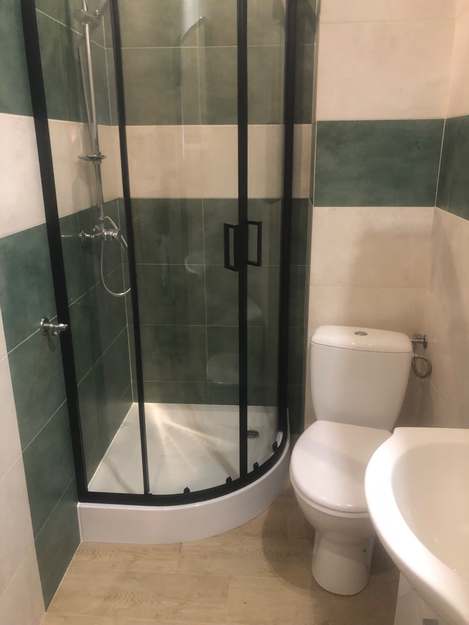 Ukończona łazienka z kabiną prysznicową z czarnymi ramami, toaletą i umywalką, wykończona płytkami w pasy zieleni i bieli.