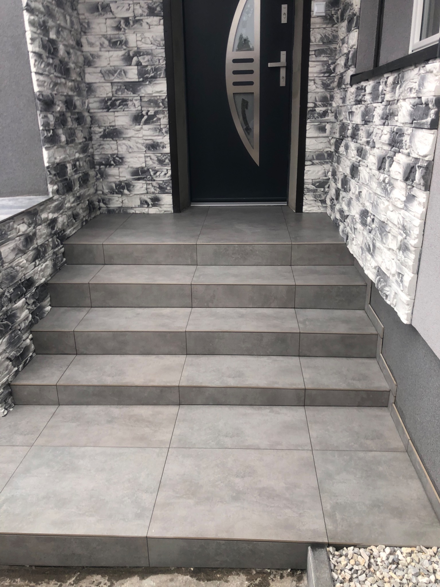 Zewnętrzne schody wejściowe wyłożone szarą, matową płytką ceramiczną, prowadzące do czarnych drzwi z metalowymi zdobieniami, ściany wykończone dekoracyjną okładziną imitującą kamień.