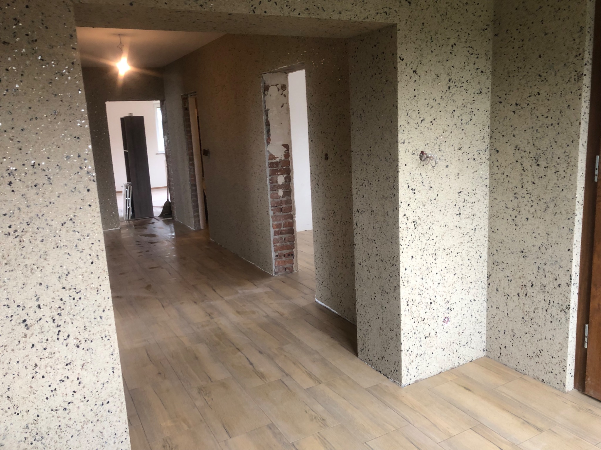 Korytarz w trakcie remontu z wykończonymi ścianami w dekoracyjnym tynku z drobnymi kamyczkami, drewnopodobną podłogą i widocznym fragmentem ściany z cegły w przejściu.