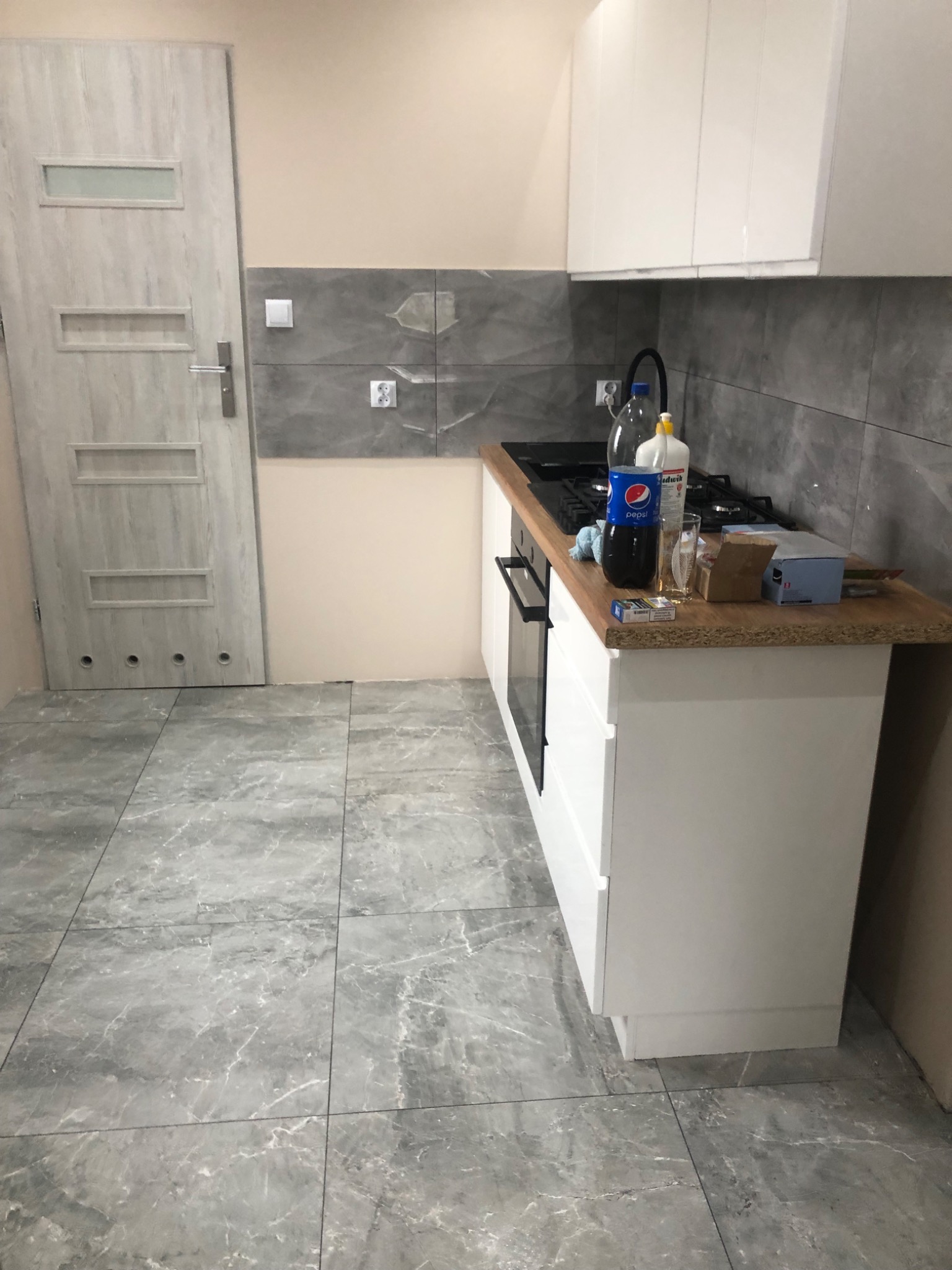 Wykończona kuchnia z szarymi płytkami podłogowymi imitującymi marmur, białymi szafkami kuchennymi i szarymi płytkami ściennymi; na blacie widoczne butelki, naczynia i paczka papierosów.