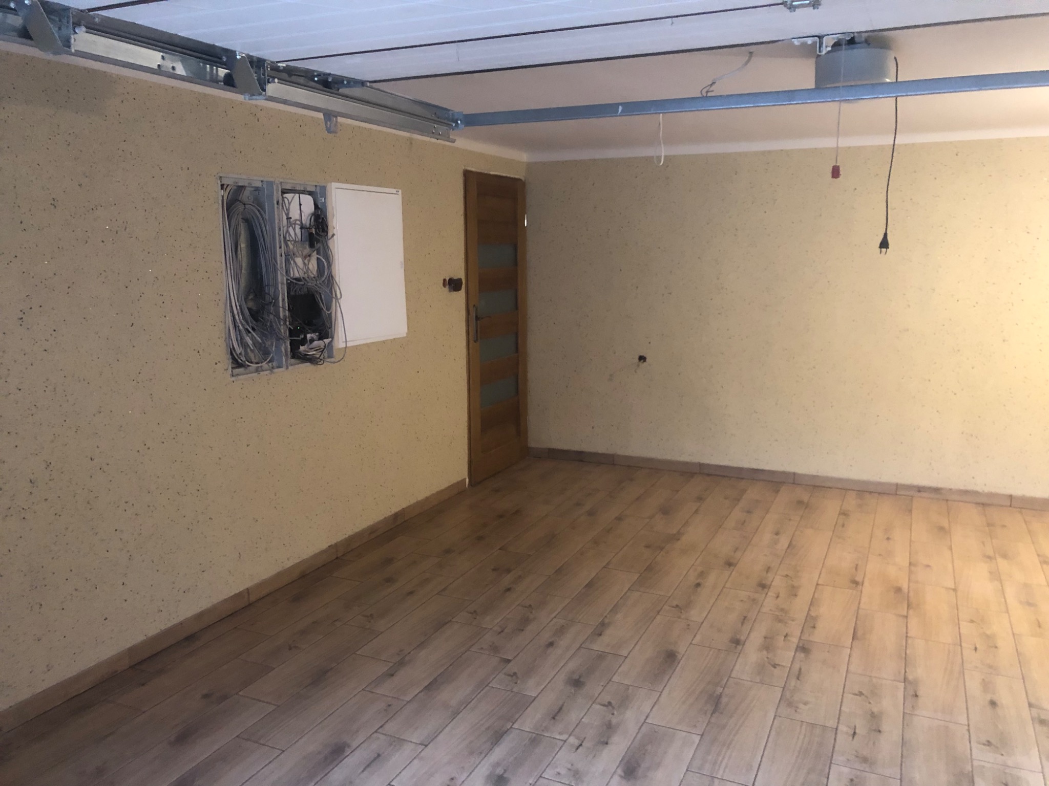Wykończone wnętrze garażu z położoną podłogą z paneli imitujących drewno, widoczne kable wystające ze ściany oraz szyny do bramy garażowej pod sufitem.