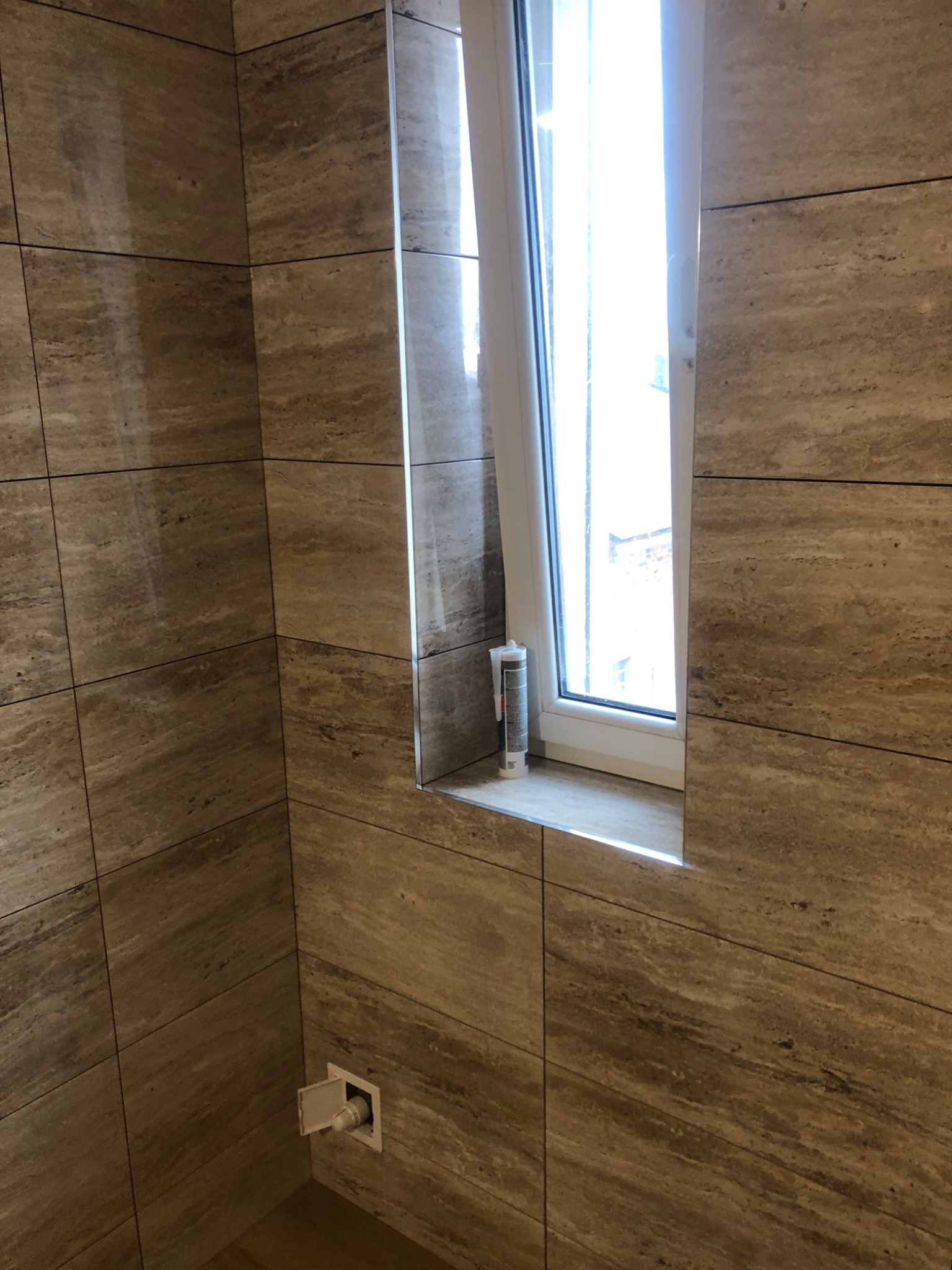 Wykończona ściana w łazience z płytek imitujących trawertyn, okno z białymi ramami i tubka silikonu na parapecie, widoczny fragment instalacji wodnej.