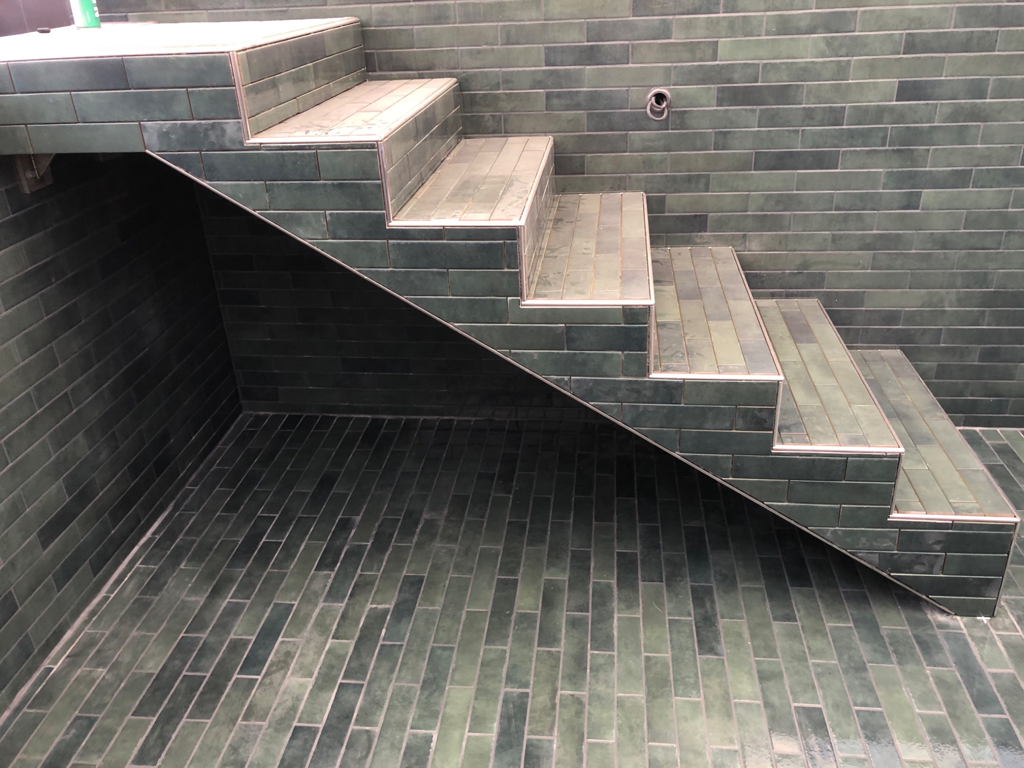 Schody wyłożone podłużnymi, zielonymi płytkami ceramicznymi z jasnymi fugami i metalowym wykończeniem krawędzi, prowadzące w dół, widoczne z bliskiej perspektywy.