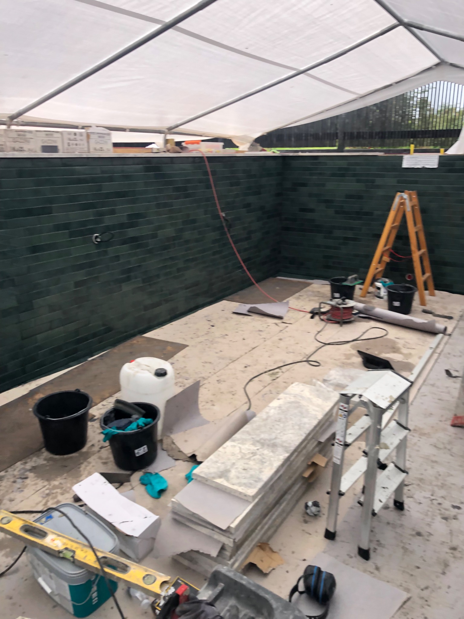 Wnętrze basenu w trakcie wykańczania: ściany wyłożone ciemnozieloną, błyszczącą glazurą, podłoga przygotowana do położenia płytek, widoczne narzędzia, drabiny i materiały budowlane pod białym...