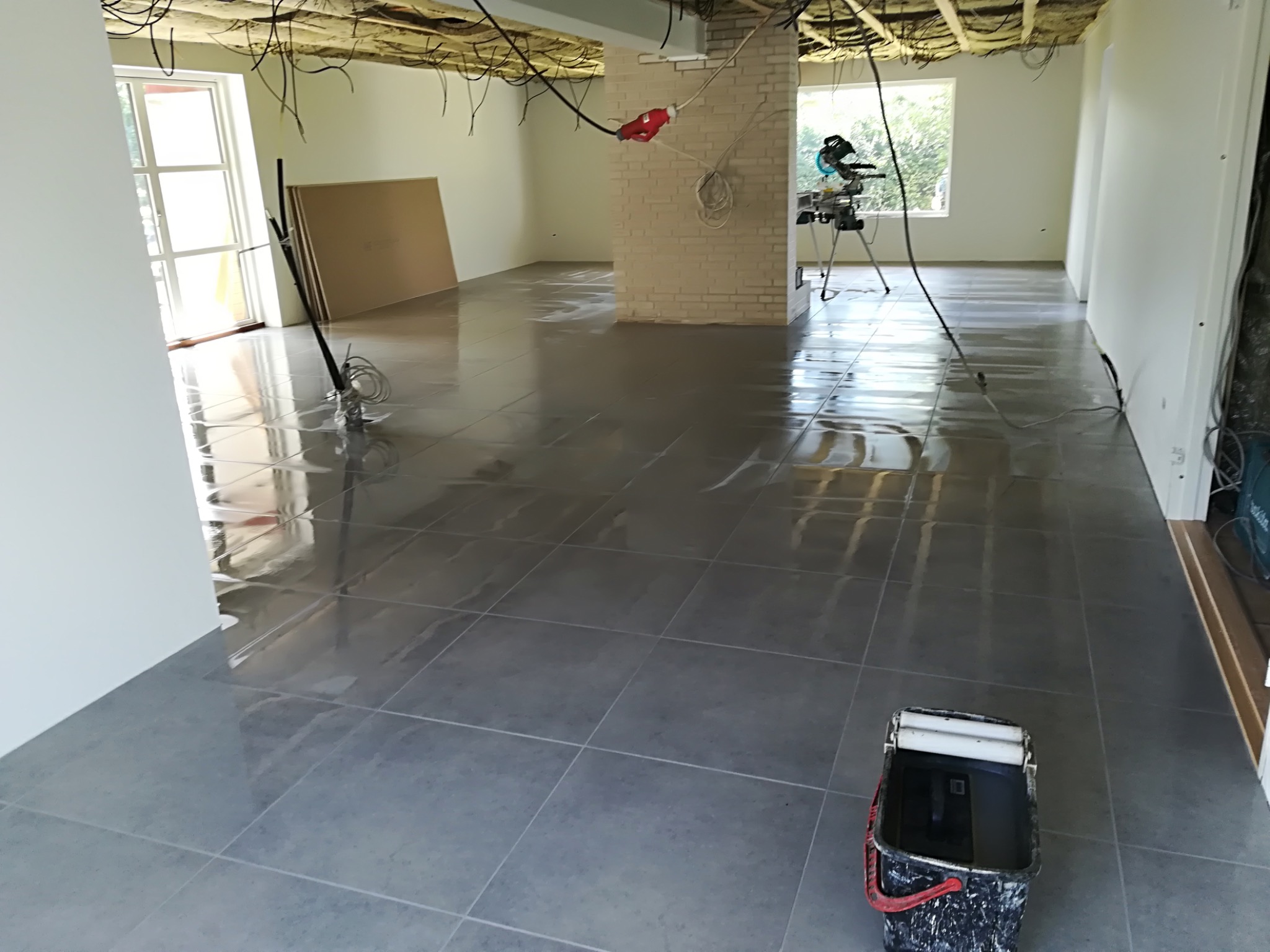 Świeżo położona, błyszcząca, szara glazura podłogowa w przestronnym, surowym wnętrzu z odsłoniętą instalacją na suficie i narzędziami glazurniczymi, w tym wiadro z rolką do czyszczenia.