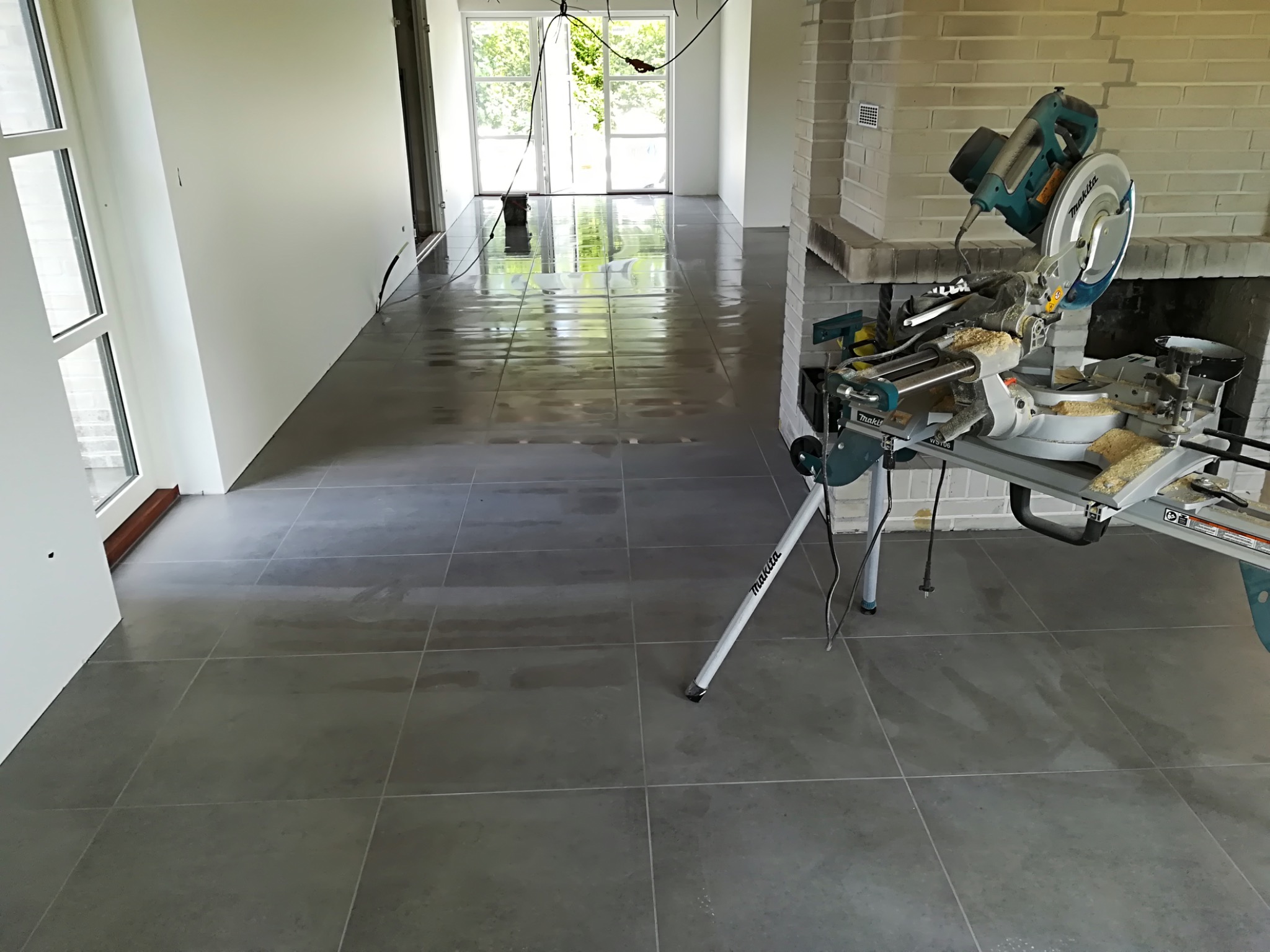 Ułożone szare płytki podłogowe w jasnym pomieszczeniu, widoczna piła stołowa Makita z resztkami ciętego materiału, ślady po wodzie na płytkach.