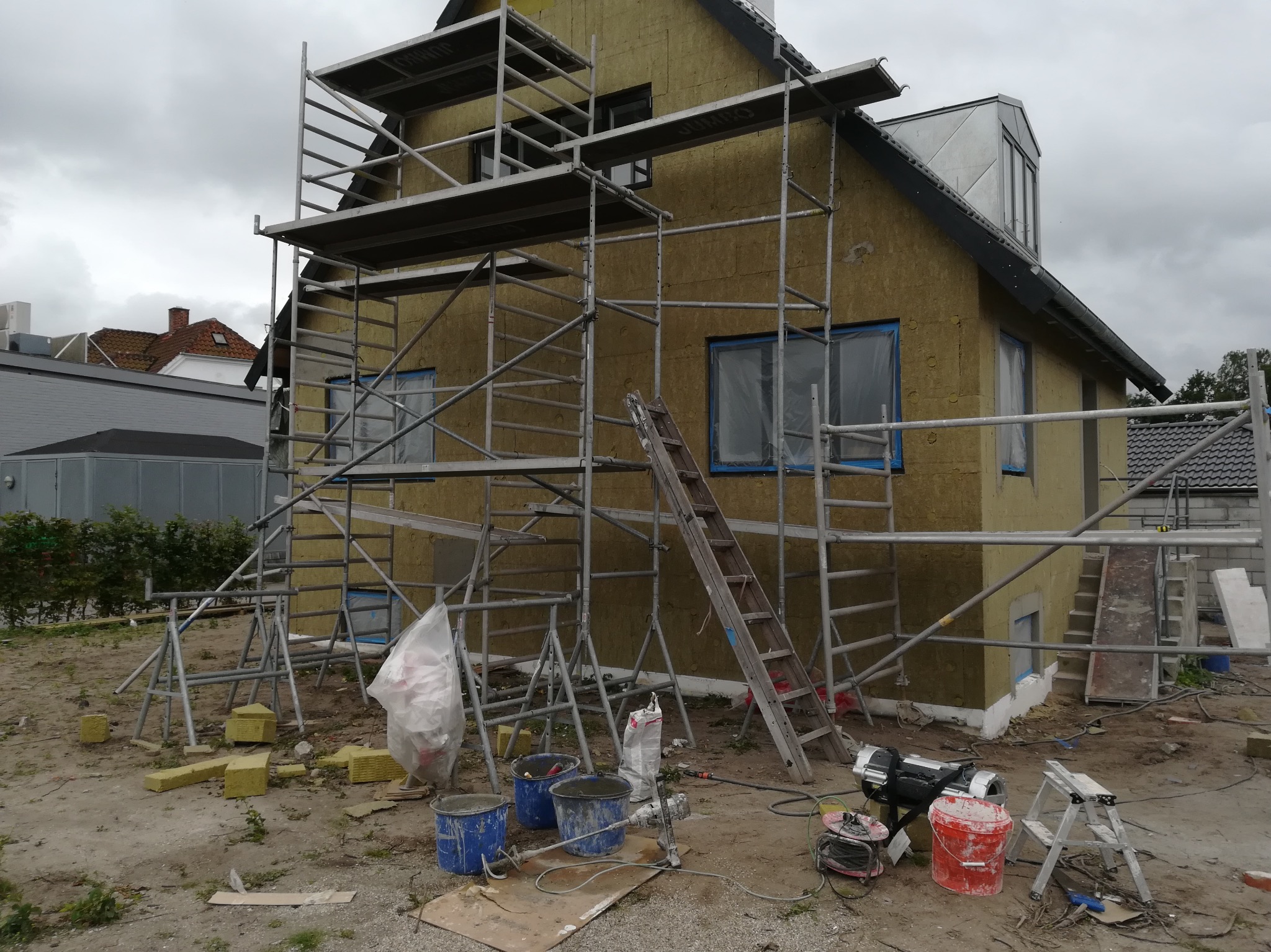 Dom w trakcie ocieplania, ściany pokryte żółtą izolacją, rusztowania dookoła, widoczne narzędzia i materiały budowlane, pochmurne niebo.