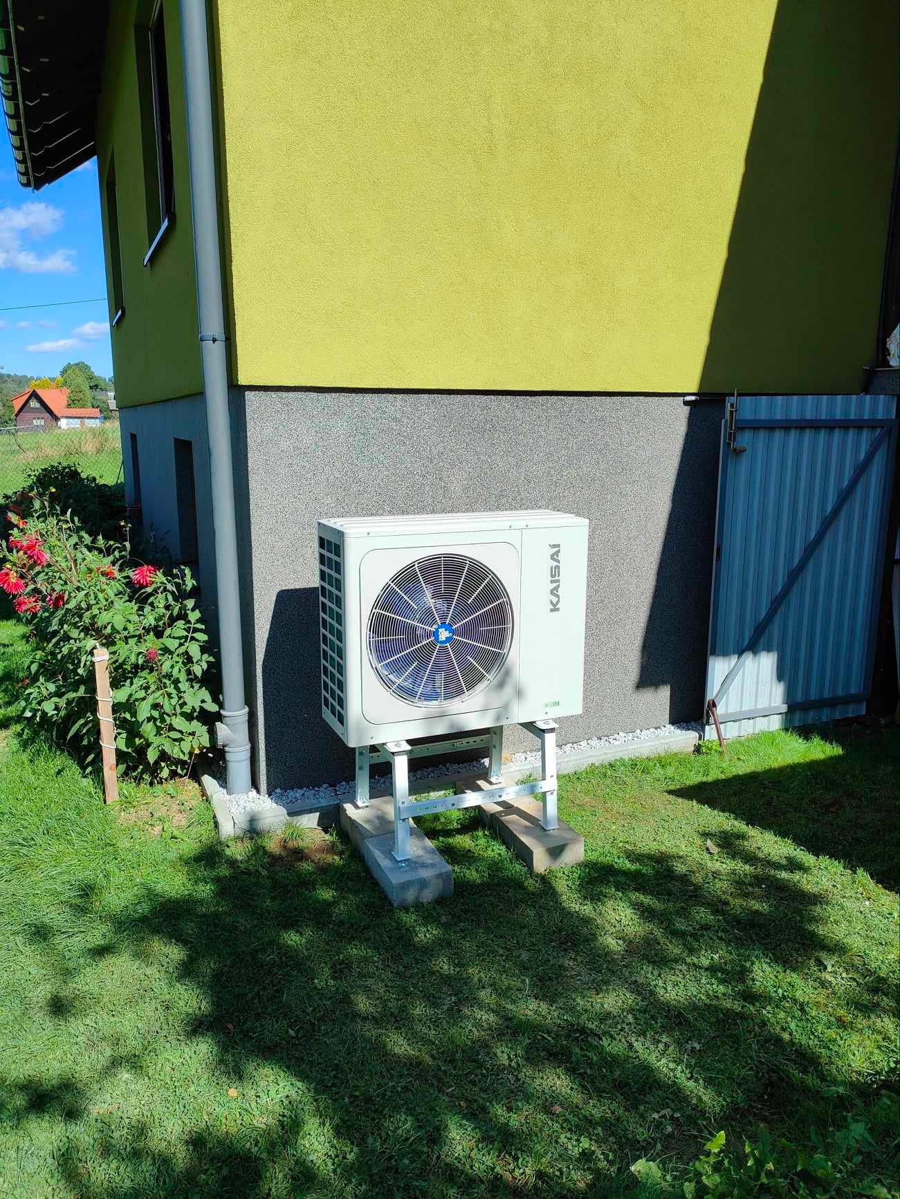 Zewnętrzna jednostka pompy ciepła Kaisai zamontowana na betonowych podstawach przy budynku z żółtą elewacją, widoczne kwiaty i fragment metalowej bramy.