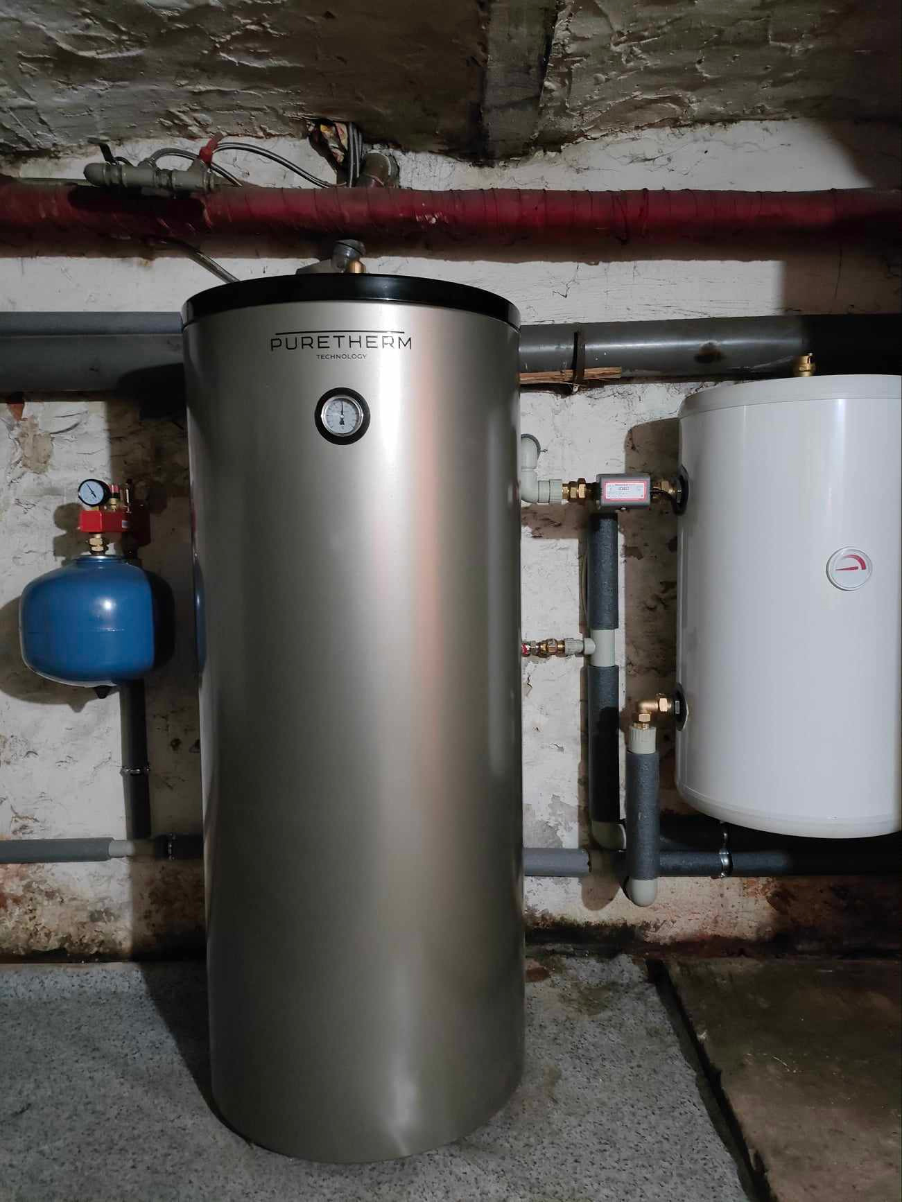 Nowoczesny, srebrny zbiornik buforowy Puretherm z manometrem, podłączony do instalacji grzewczej w piwnicy o surowych ścianach, obok biały zasobnik ciepłej wody i niebieskie naczynie przeponowe.