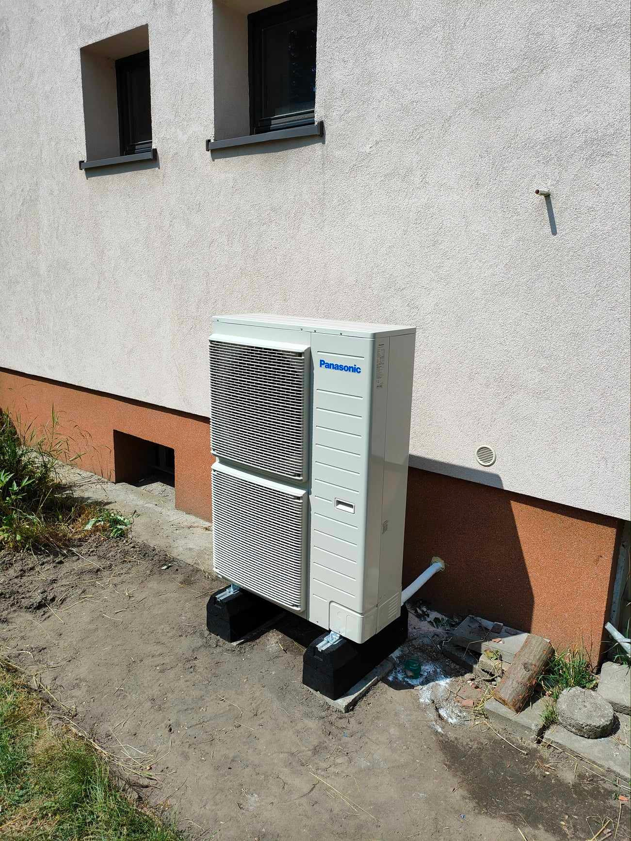 Zewnętrzna jednostka pompy ciepła Panasonic zamontowana na czarnych podstawach przy ścianie budynku z widocznym otworem wentylacyjnym i rurą odprowadzającą skropliny.