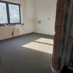 Świeżo wylana, gładka posadzka w jasnym pomieszczeniu z widocznymi instalacjami elektrycznymi w ścianie i fragmentem surowego muru z cegły.