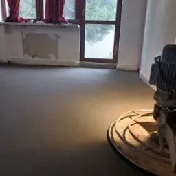 Wnętrze pokoju z świeżo wylaną, gładką posadzką, obok widoczna zacieraczka do betonu, w tle okno z czerwonymi zasłonami.