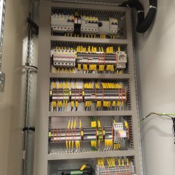 Instal volt - Rozdzielnia elektryczna w szarej obudowie, widoczne liczne przewody w żółtych oznaczeniach, zabezpieczenia, listwy zaciskowe i gniazdo elektryczne.