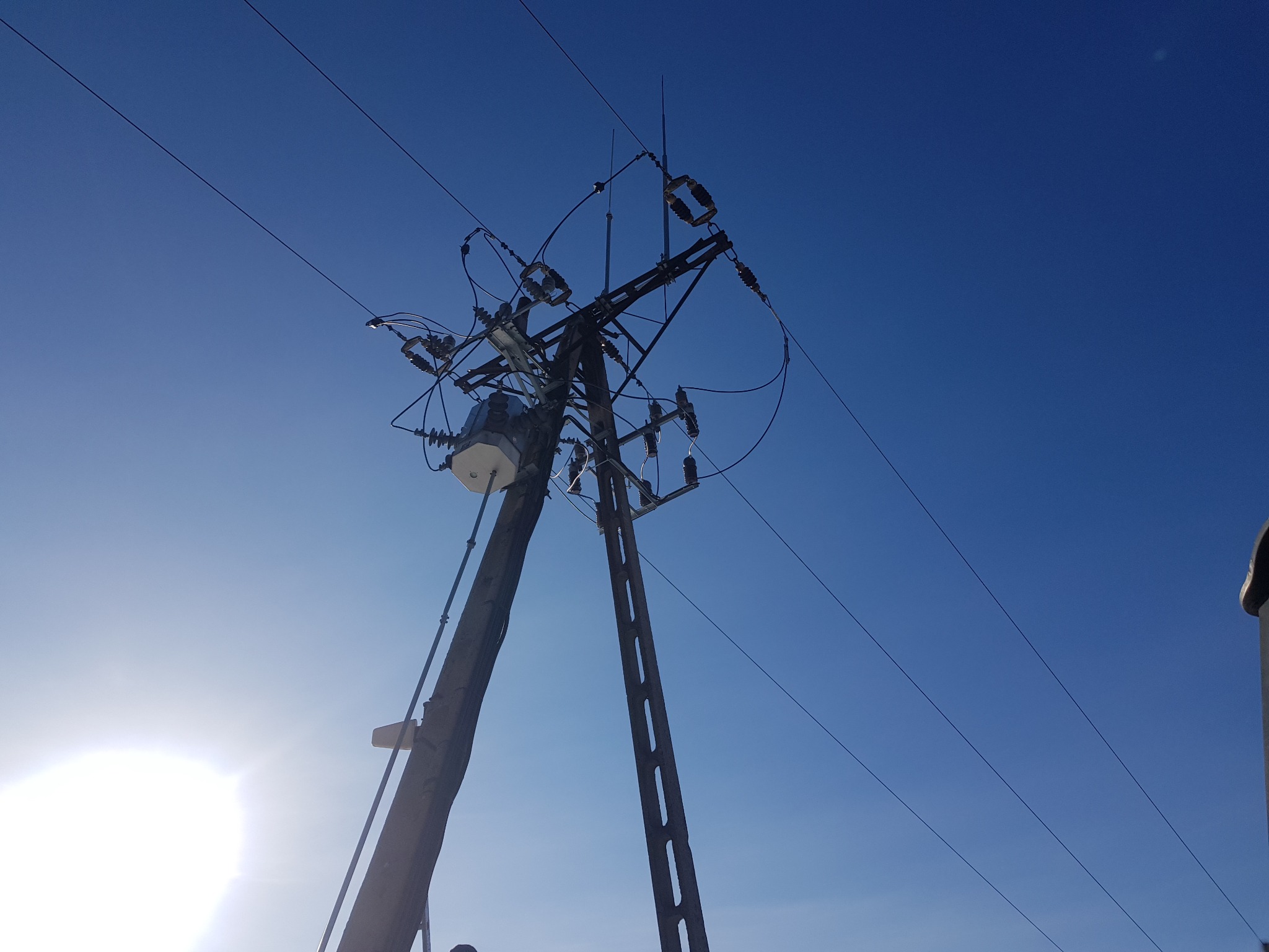 Słup energetyczny z transformatorem i liniami wysokiego napięcia, sfotografowany pod kątem w słoneczny dzień na tle błękitnego nieba.