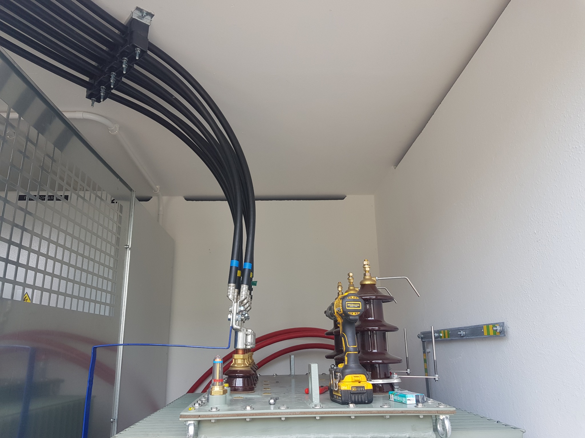 Transformator w białym pomieszczeniu z podłączonymi grubymi kablami zasilającymi, widoczny wkrętak akumulatorowy DeWalt na transformatorze.