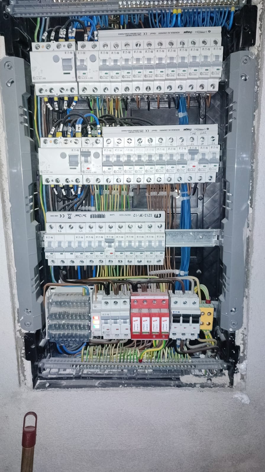 Rozdzielnica elektryczna z licznymi zabezpieczeniami i przewodami w różnych kolorach, zamontowana w ścianie, widoczne ślady po montażu i pył.