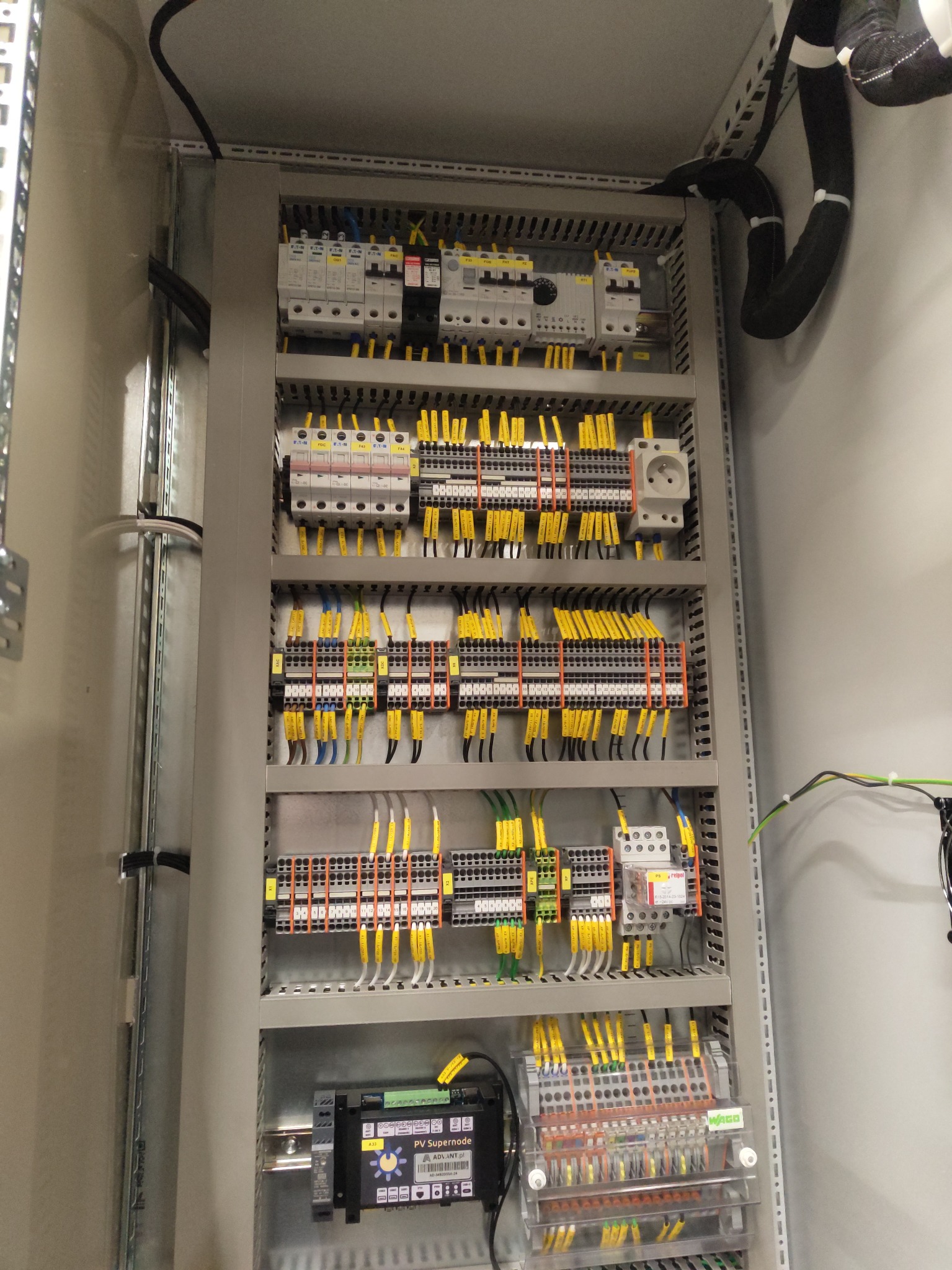 Rozdzielnia elektryczna w szarej obudowie, widoczne liczne przewody w żółtych oznaczeniach, zabezpieczenia, listwy zaciskowe i gniazdo elektryczne.