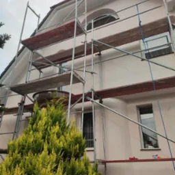 Dom w Szydłowcu z jasnoróżową elewacją, w trakcie prac na rusztowaniu, widoczne narzędzia i materiały malarskie na podeście rusztowania, iglak w pierwszym planie.