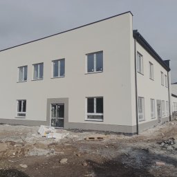 Usługi Ogólnobudowlane - Nowo otynkowany, dwukondygnacyjny budynek biurowy z białymi ścianami i szarym cokołem, z widocznymi oknami z folią ochronną i materiałami budowlanymi na placu budowy.