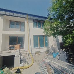 Usługi Ogólnobudowlane - Dwupiętrowy dom z żółtym wężem do ocieplania poddasza, widoczne rusztowania i materiały budowlane, częściowo otynkowana elewacja.