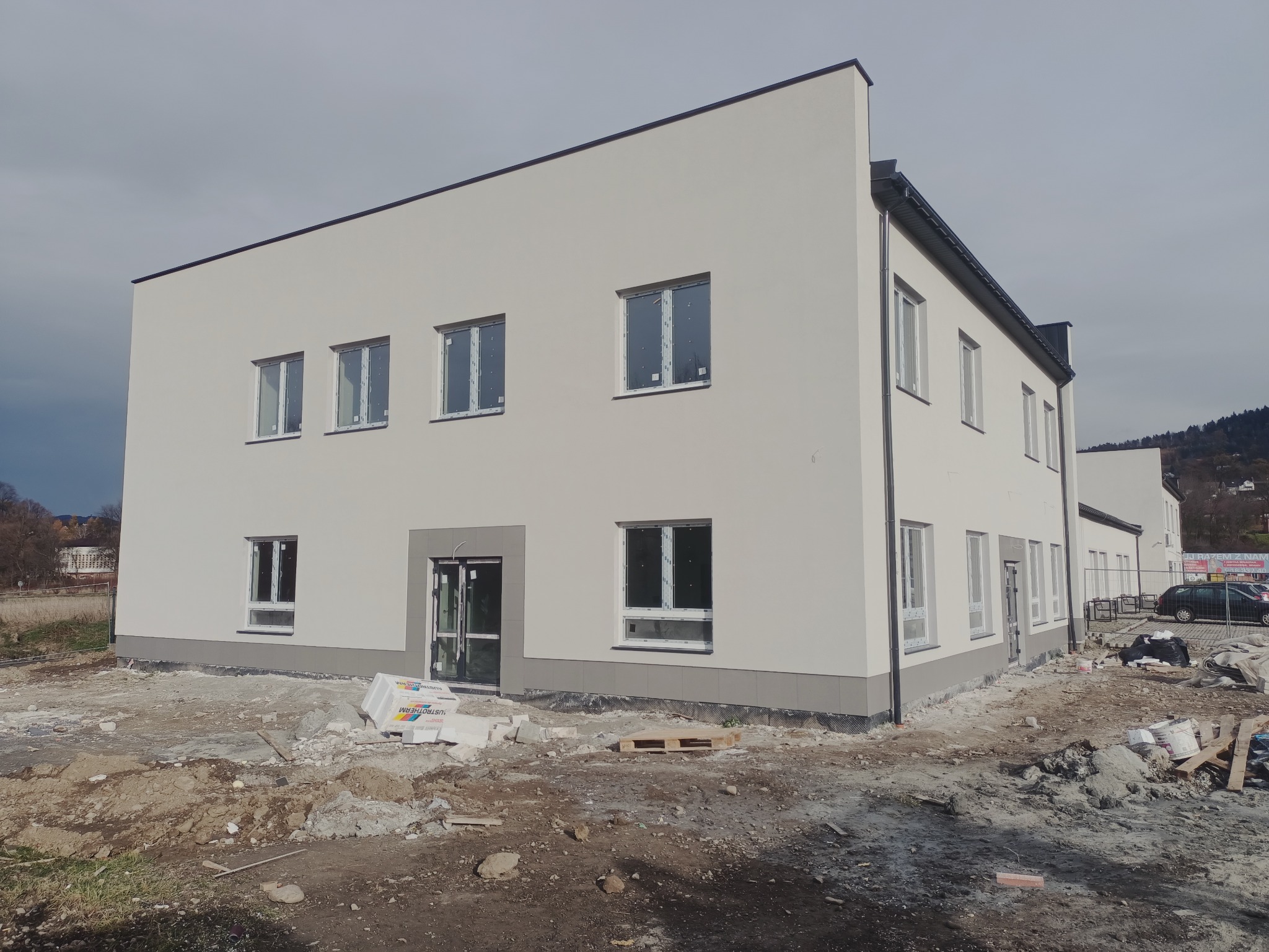 Nowo otynkowany, dwukondygnacyjny budynek biurowy z białymi ścianami i szarym cokołem, z widocznymi oknami z folią ochronną i materiałami budowlanymi na placu budowy.