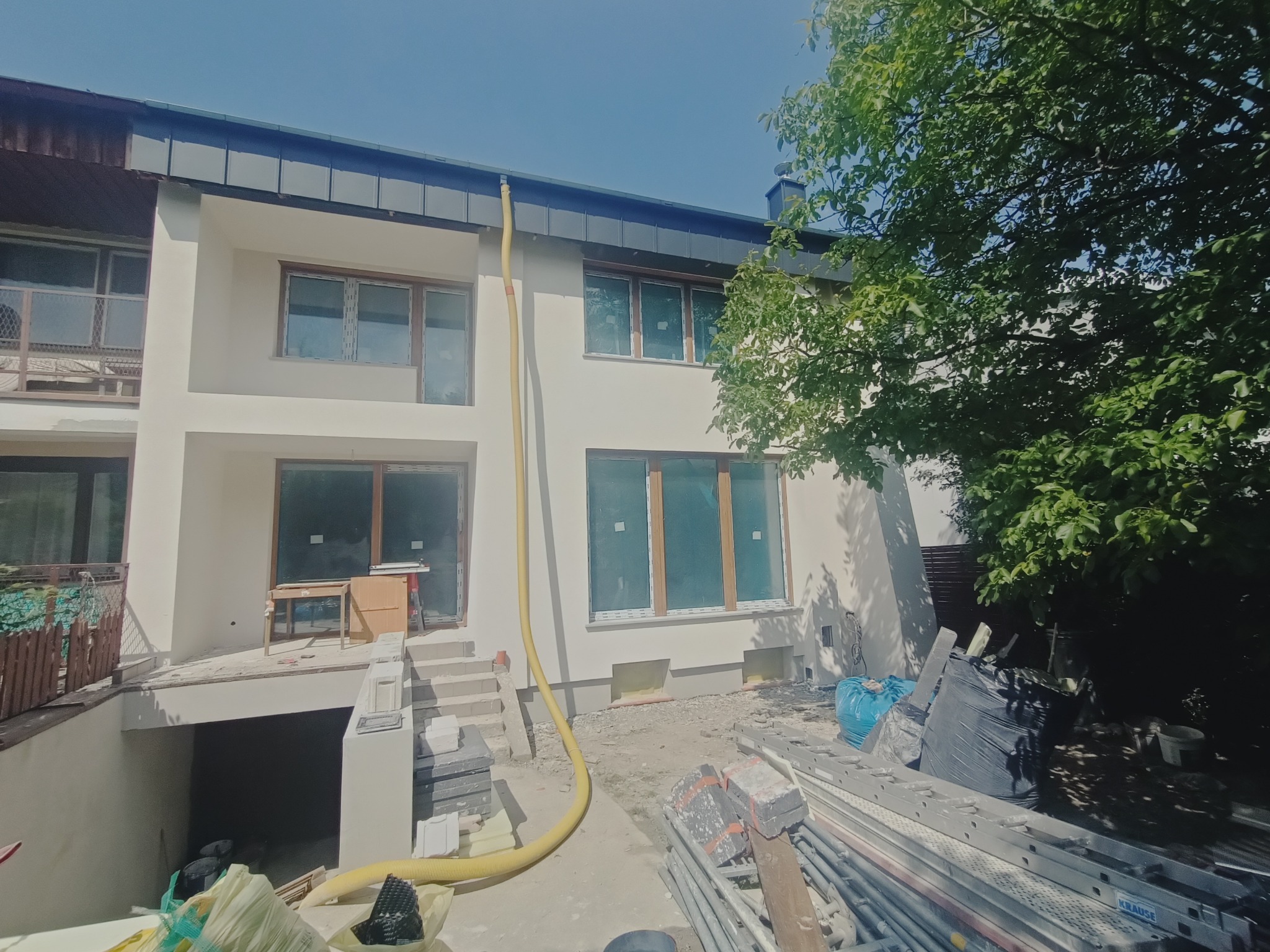 Dwupiętrowy dom z żółtym wężem do ocieplania poddasza, widoczne rusztowania i materiały budowlane, częściowo otynkowana elewacja.