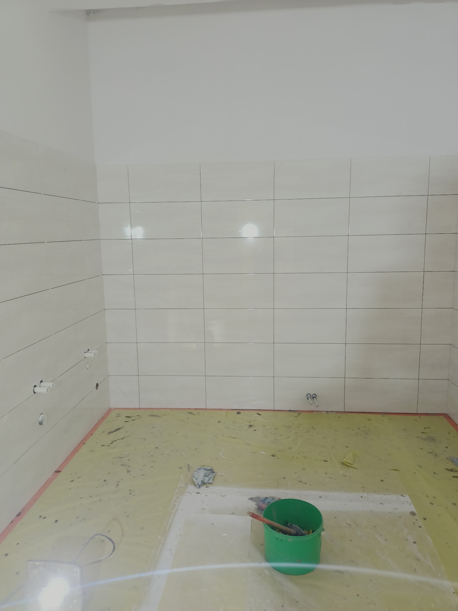 Świeżo wykafelkowana ściana w łazience z widocznymi przyłączami wodno-kanalizacyjnymi, podłoga zabezpieczona folią malarską, zielone wiadro z narzędziami na posadzce.