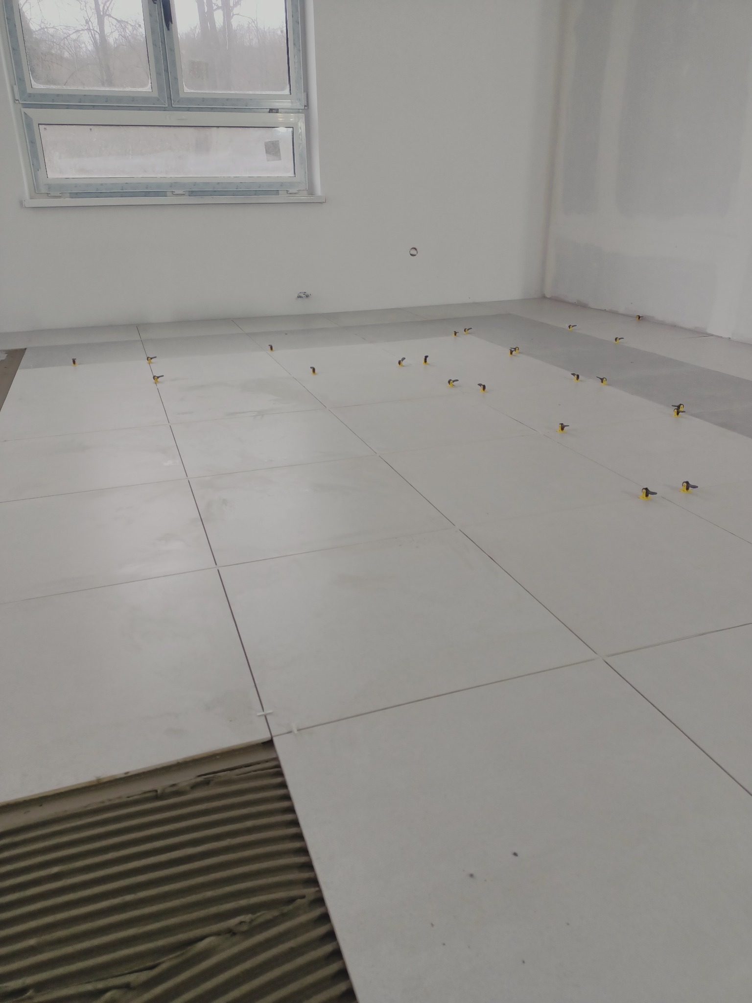 Jasnoszare płytki podłogowe w trakcie układania, widoczna warstwa kleju z charakterystycznymi rowkami i żółte krzyżyki dystansowe, surowe wnętrze w trakcie remontu.
