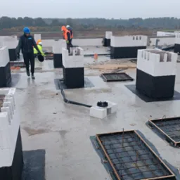 Prace na dachu płaskim: robotnicy w kaskach, białe bloczki fundamentowe z czarną izolacją, zbrojenia w drewnianych szalunkach, system odprowadzania wody. Widok na horyzont z drzewami i budynkami.