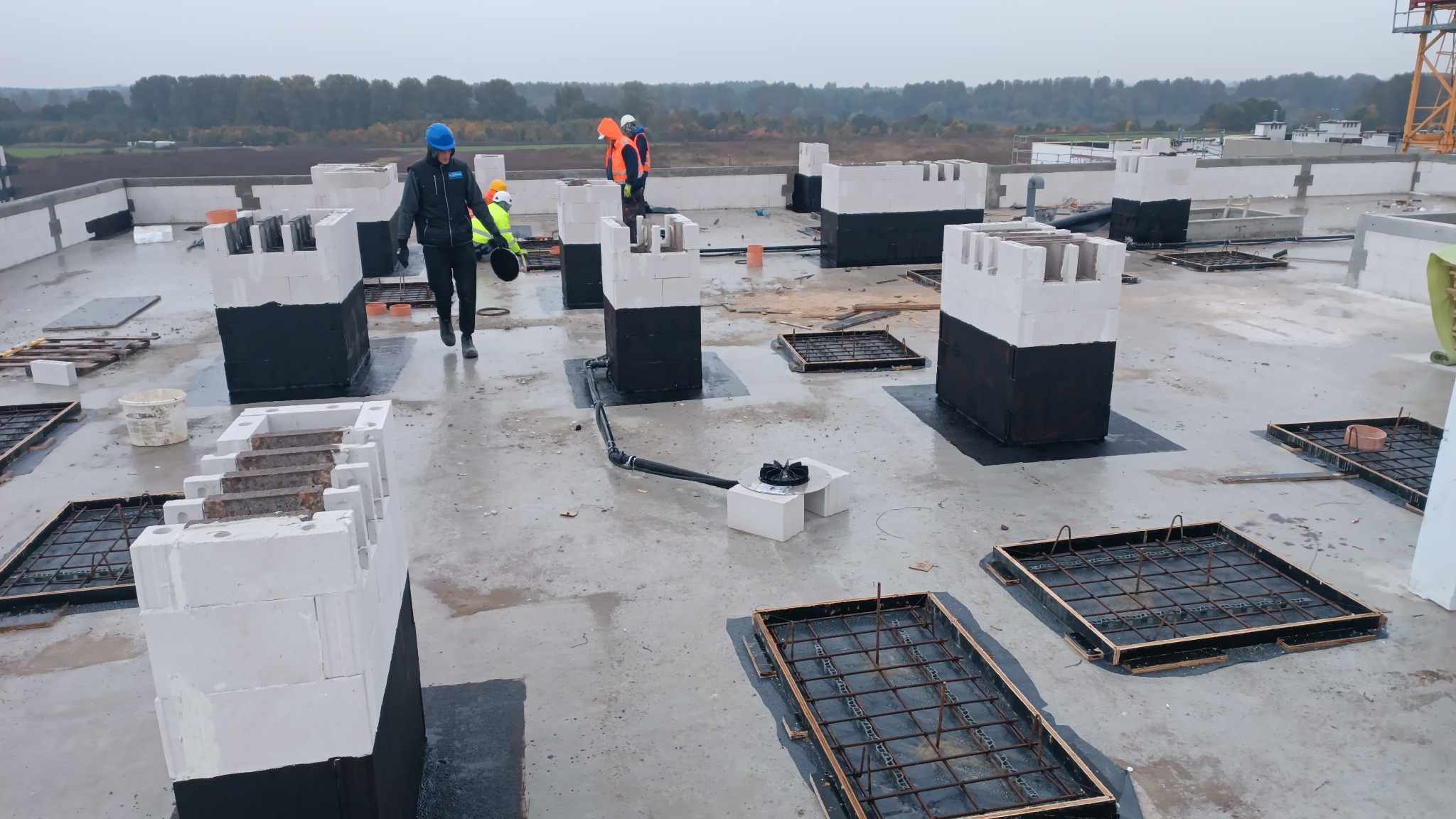 Prace na dachu płaskim: robotnicy w kaskach, białe bloczki fundamentowe z czarną izolacją, zbrojenia w drewnianych szalunkach, system odprowadzania wody. Widok na horyzont z drzewami i budynkami.