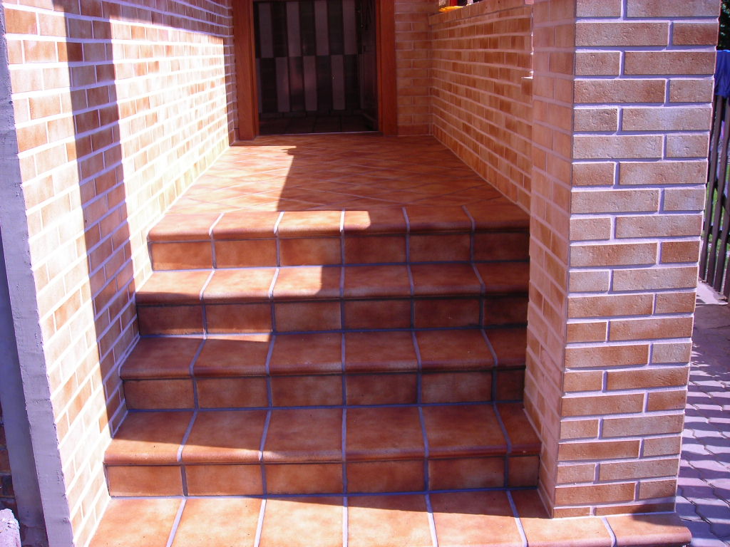 Wykończone schody wejściowe z terakoty, otoczone ceglaną ścianą i filarem, w ciepłym oświetleniu słonecznym.