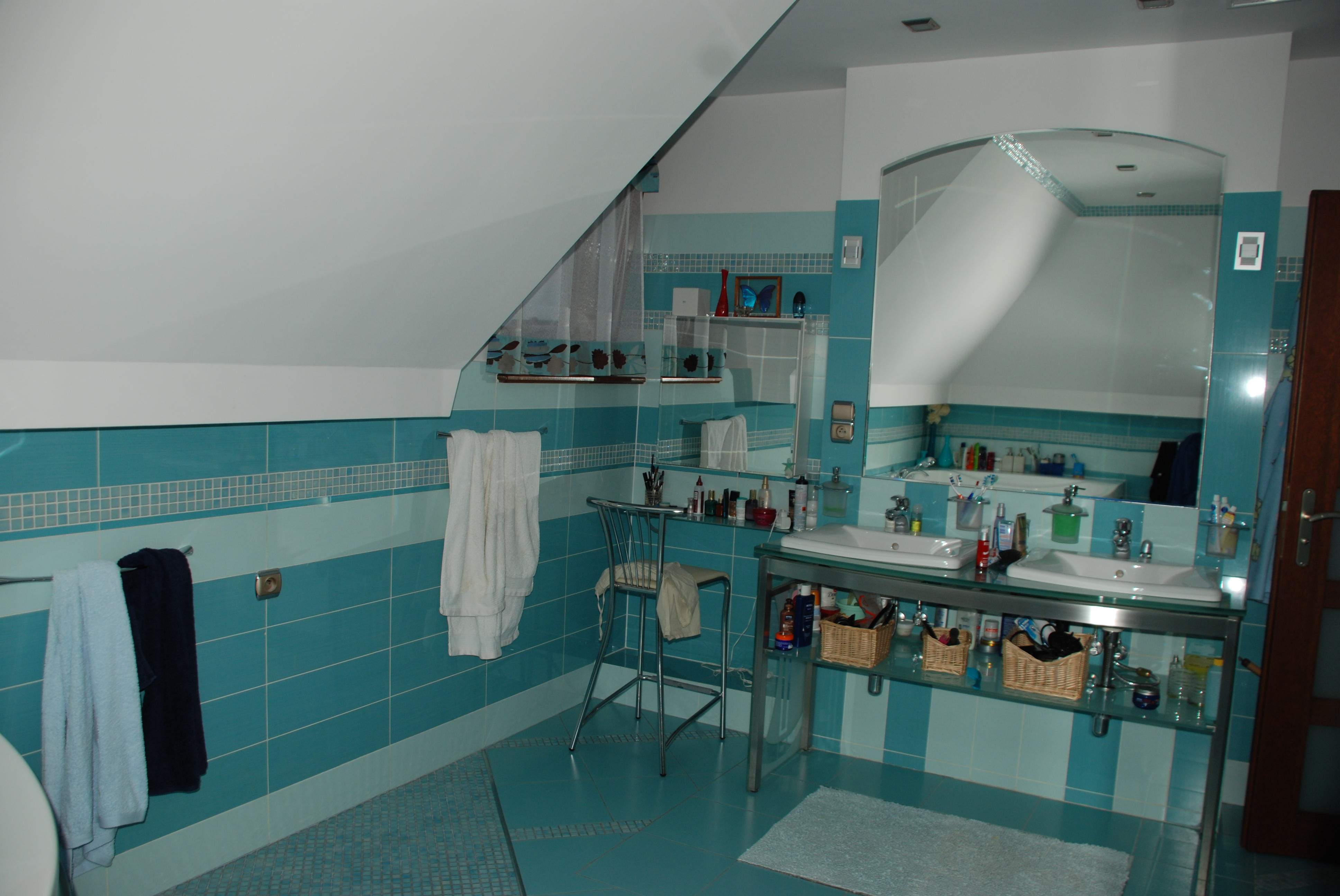 Turkusowa łazienka na poddaszu z dwiema umywalkami na szklanym blacie, lustrem z zaokrąglonymi rogami, ręcznikami i dekoracjami na półkach.