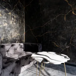 Elegancki salon z ciemnymi ścianami zdobionymi złotymi żyłkami, szarym, pikowanym welurowym narożnikiem i stolikiem z blatem imitującym marmur na złotych nóżkach.