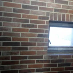 Ściana z cegły klinkierowej w odcieniach brązu i czerwieni, z wbudowanym oknem PCV z widoczną ramą. Cegły ułożone w regularny wzór z jasną fugą.